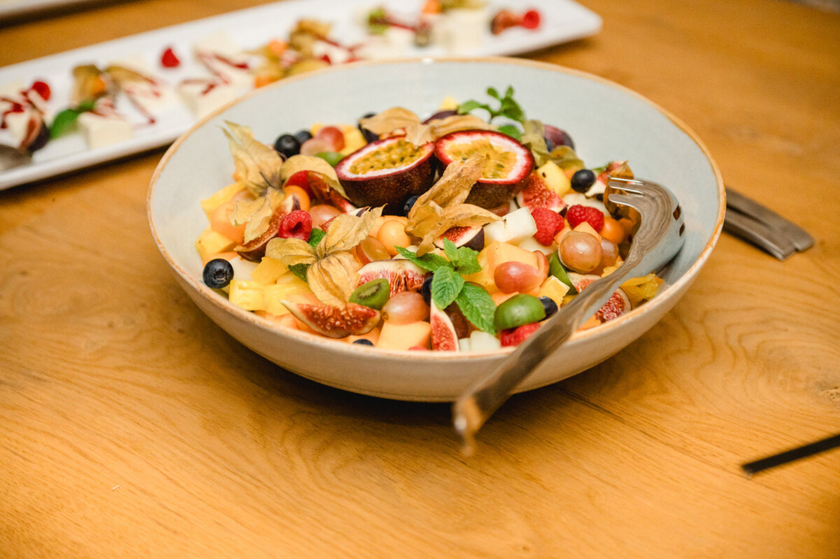 Eine Schüssel Obstsalat mit Passionsfrüchten, Feigen, Physalis, Blaubeeren und Melonenstücken auf einem Holztisch, in der Schüssel liegt eine Gabel.
