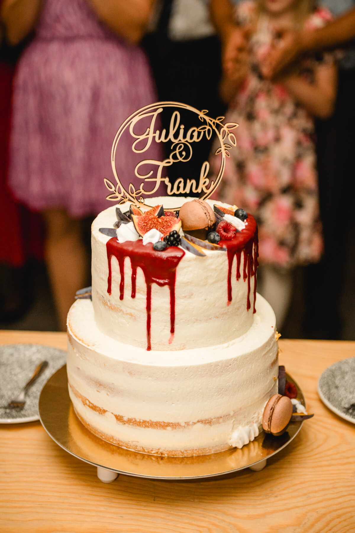 Zweistöckige weiße Torte mit rotem Guss, Feigen und Macarons. „Julia & Frank“-Dekoration. Menschen im Hintergrund.