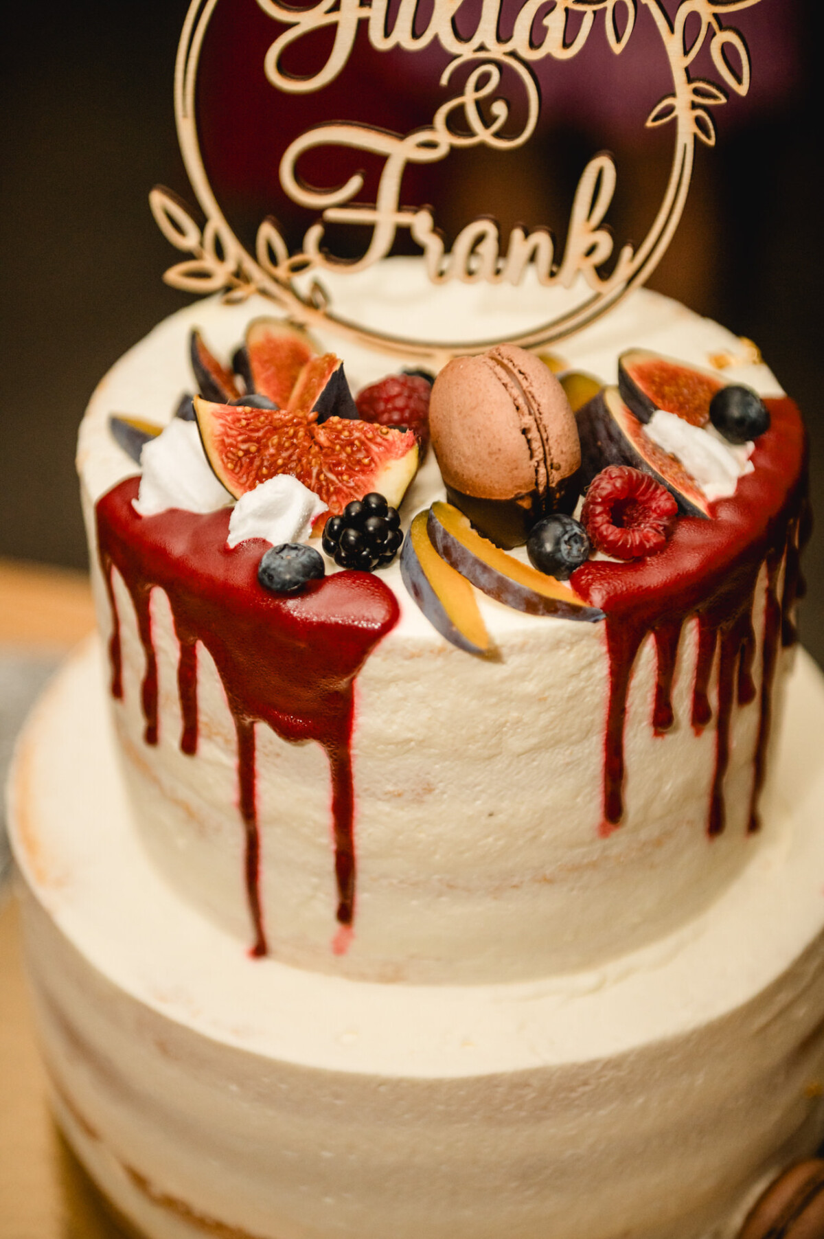 Zweistöckiger weißer Kuchen mit rotem Zuckerguss, garniert mit Feigen, Beeren, einem Makronenkuchen und einem dekorativen Holzaufsatz.