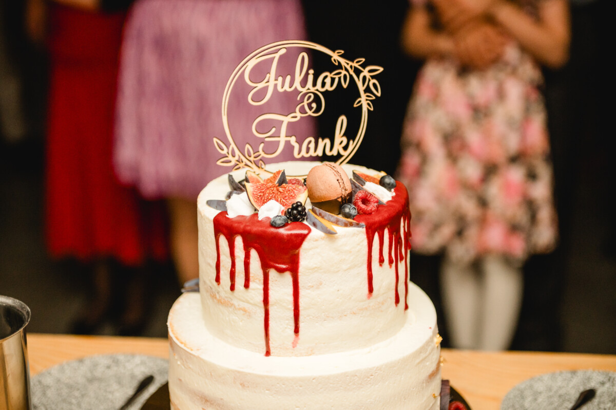 Eine zweistöckige Torte mit rotem Zuckerguss, garniert mit einem Macaron, Früchten und einem dekorativen Topper mit Namen. Im Vordergrund sind Teller zu sehen, im Hintergrund sind verschwommen Menschen in Kleidern zu sehen.