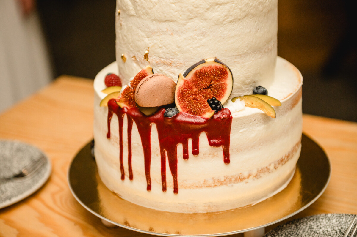 Zweistöckige Torte mit weißem Zuckerguss, garniert mit Feigen, Macarons, Beeren und einem roten Tropfen. Auf einem Holztisch mit zwei Tellern daneben.