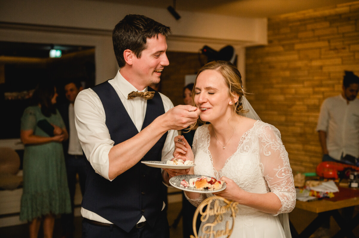 Ein Bräutigam in einer Weste füttert eine Braut in einem Spitzenkleid während einer Hochzeitsfeier mit Kuchen, beide lächeln freudig.