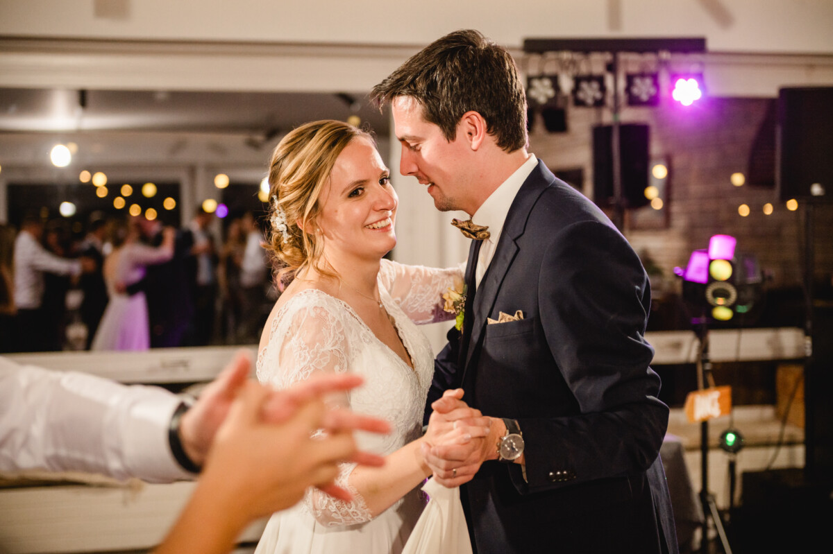 Paar in Abendgarderobe lächelt und tanzt bei einer gut beleuchteten Veranstaltung, mit anderen Gästen im Hintergrund.