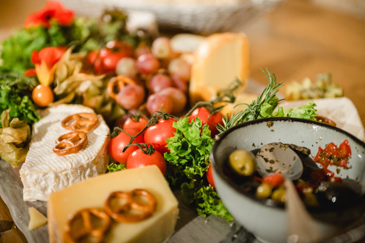 Eine Platte mit verschiedenen Käsesorten, Brezeln, Tomaten, Trauben und Blattgemüse. Daneben steht eine kleine Schüssel mit Oliven und einem Löffel.