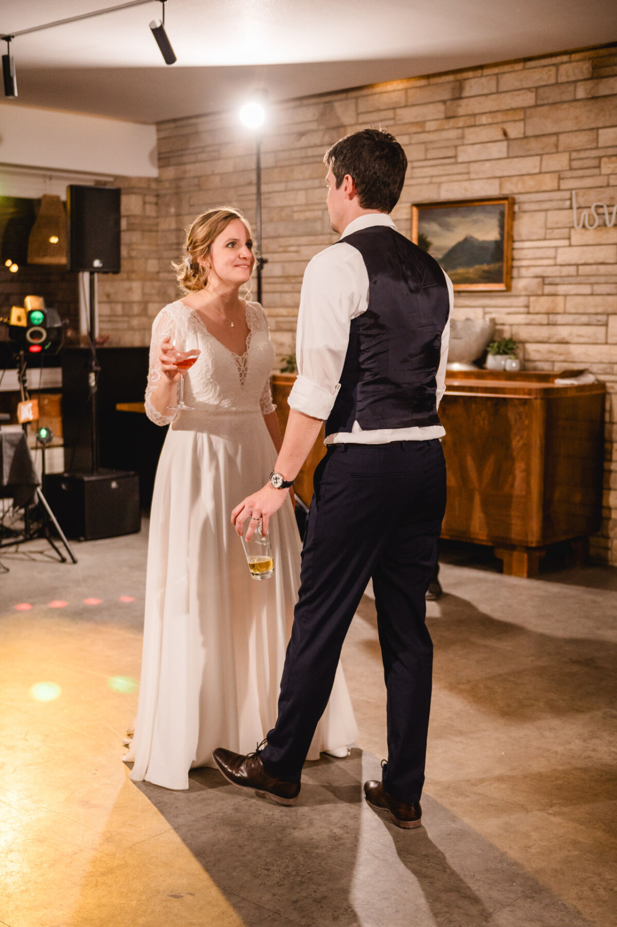 Braut und Bräutigam verbringen einen Moment auf der Tanzfläche einer Hochzeitsfeier mit Getränken in der Hand. Die Kulisse ist von einer Backsteinmauer umgeben und die Beleuchtung ist minimal.