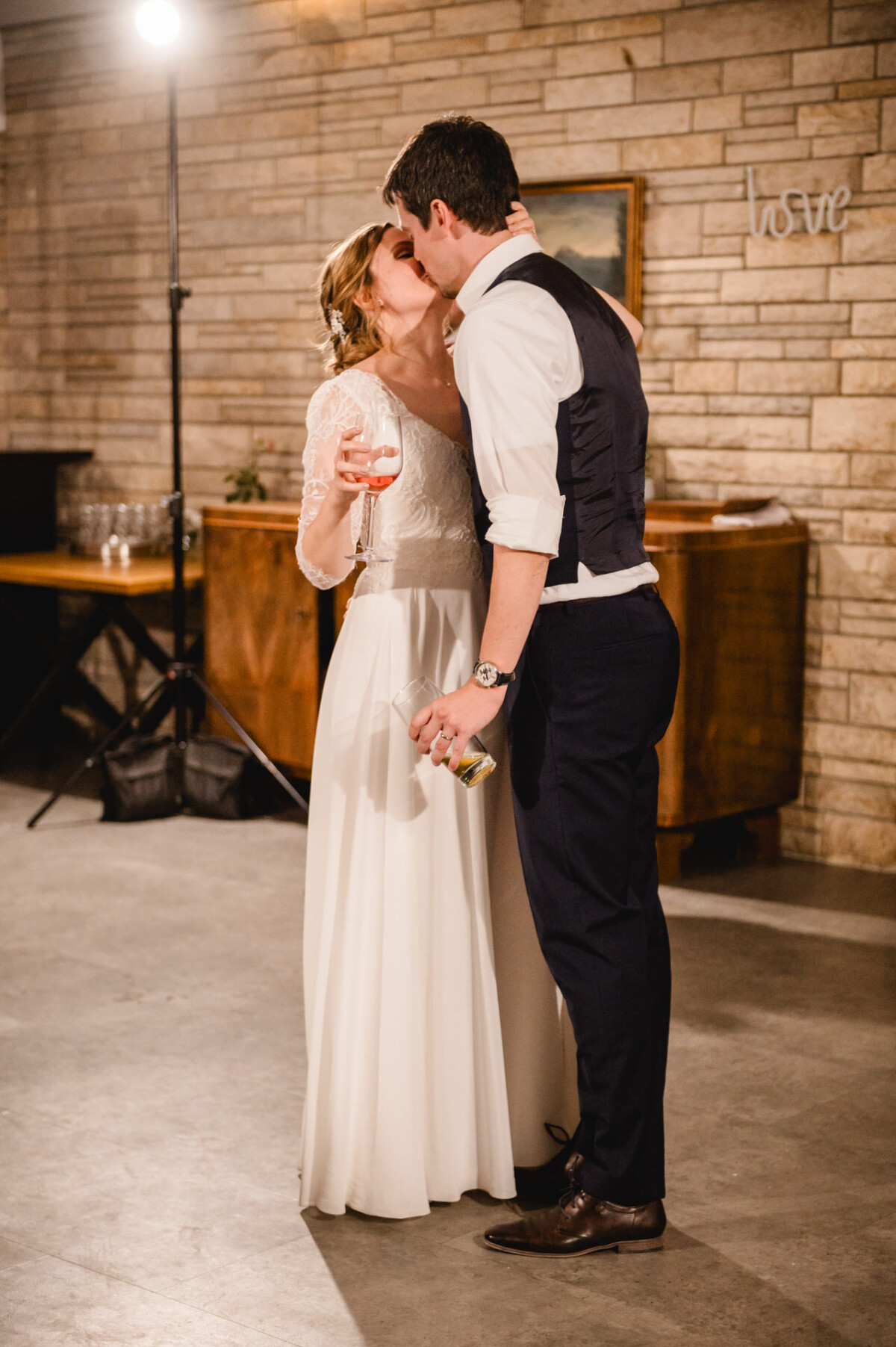 Braut und Bräutigam küssen sich und halten Getränke in der Hand. Die Braut trägt ein weißes Kleid, der Bräutigam ein weißes Hemd und eine weiße Weste. Die Szene scheint in einem Gebäude mit einer Ziegelmauer zu spielen.