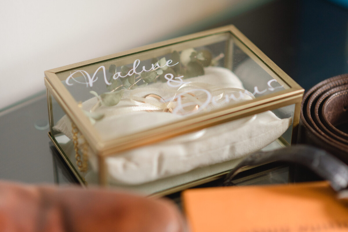 Eine gläserne Hochzeitsbox mit der Aufschrift „Nadine & Dennis“, in der sich ein weißes Kissen befindet und die auf einer Fläche neben einem Gürtel und einem Buch steht.