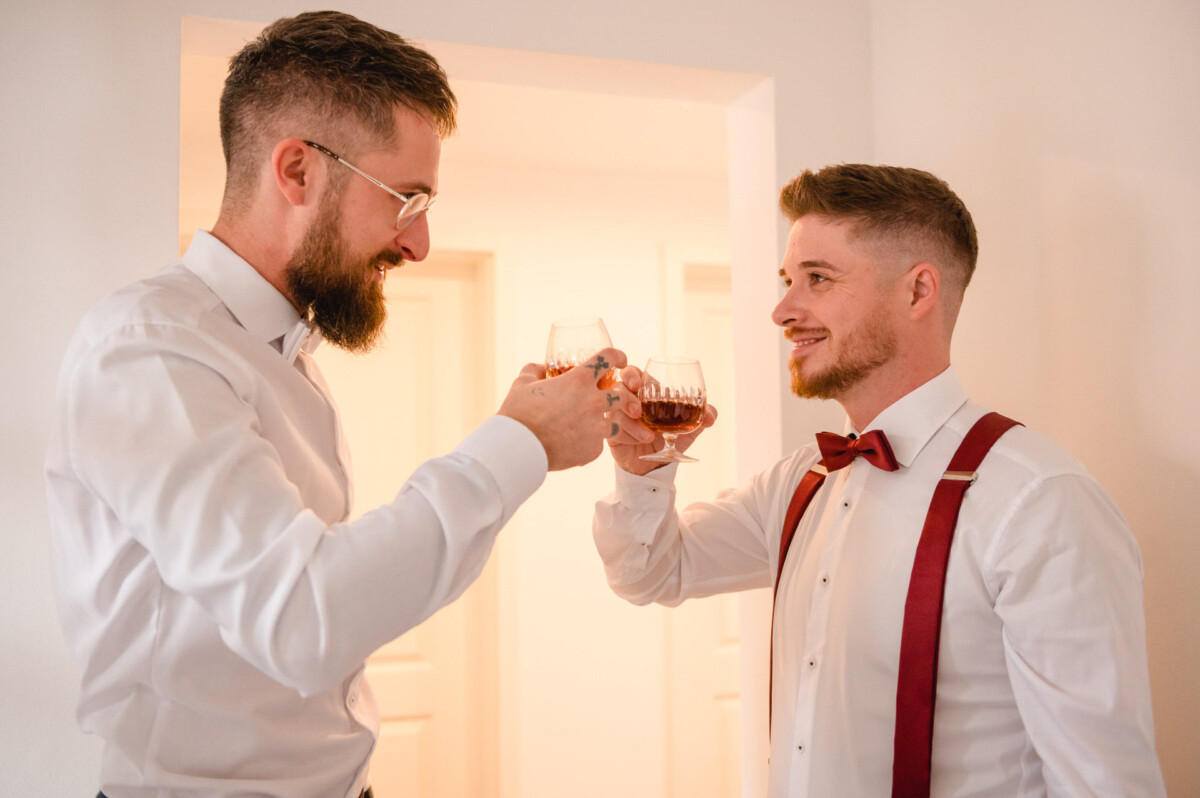 Zwei Männer in Abendgarderobe mit Hosenträgern stoßen mit Weingläsern an und lächeln sich in einem warm beleuchteten Raum an.