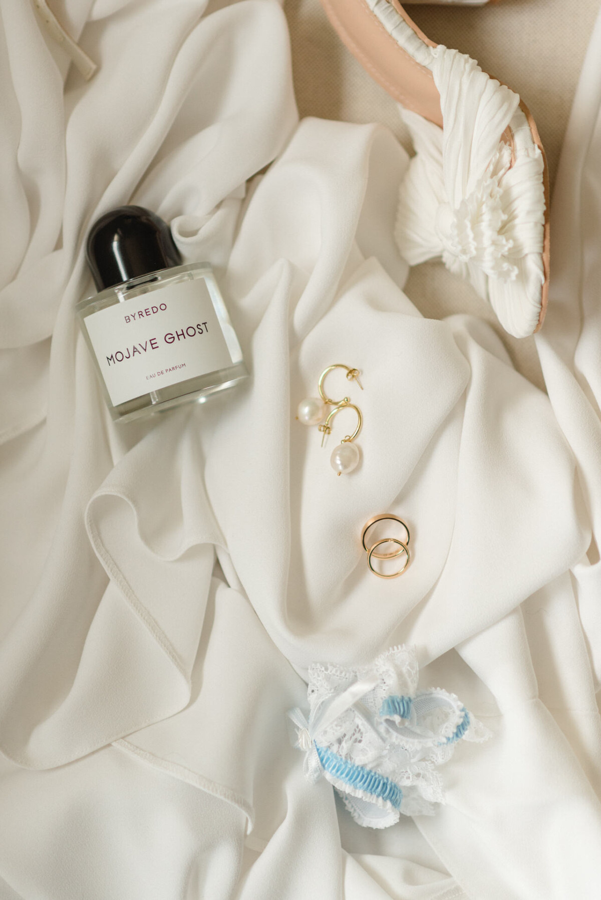 Elegante Hochzeitsaccessoires, darunter ein Parfümflakon, ein Stöckelschuh, ein Perlenohrring und Ringe auf einem drapierten weißen Stoff.