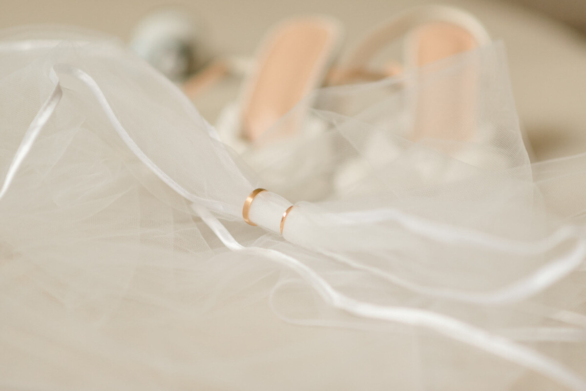 Ein goldener Ehering auf weichem Tüllstoff mit eleganten Brautschuhen, die im Hintergrund unscharf sind.