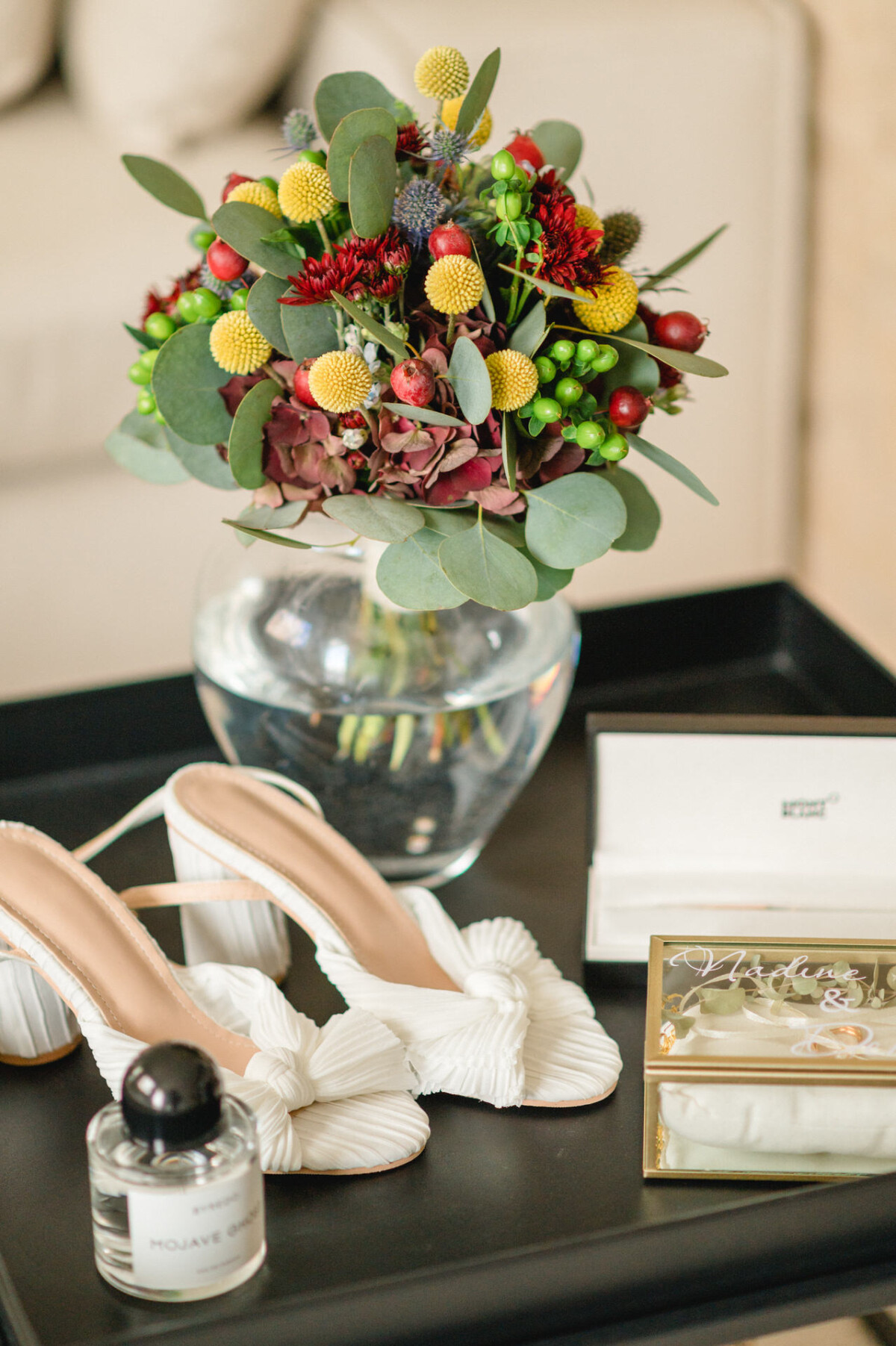 Ein farbenfroher Strauß gemischter Blumen in einer Glasvase auf einem schwarzen Tablett, begleitet von Brautschuhen, einer Schmuckschatulle und einer Parfümflasche.