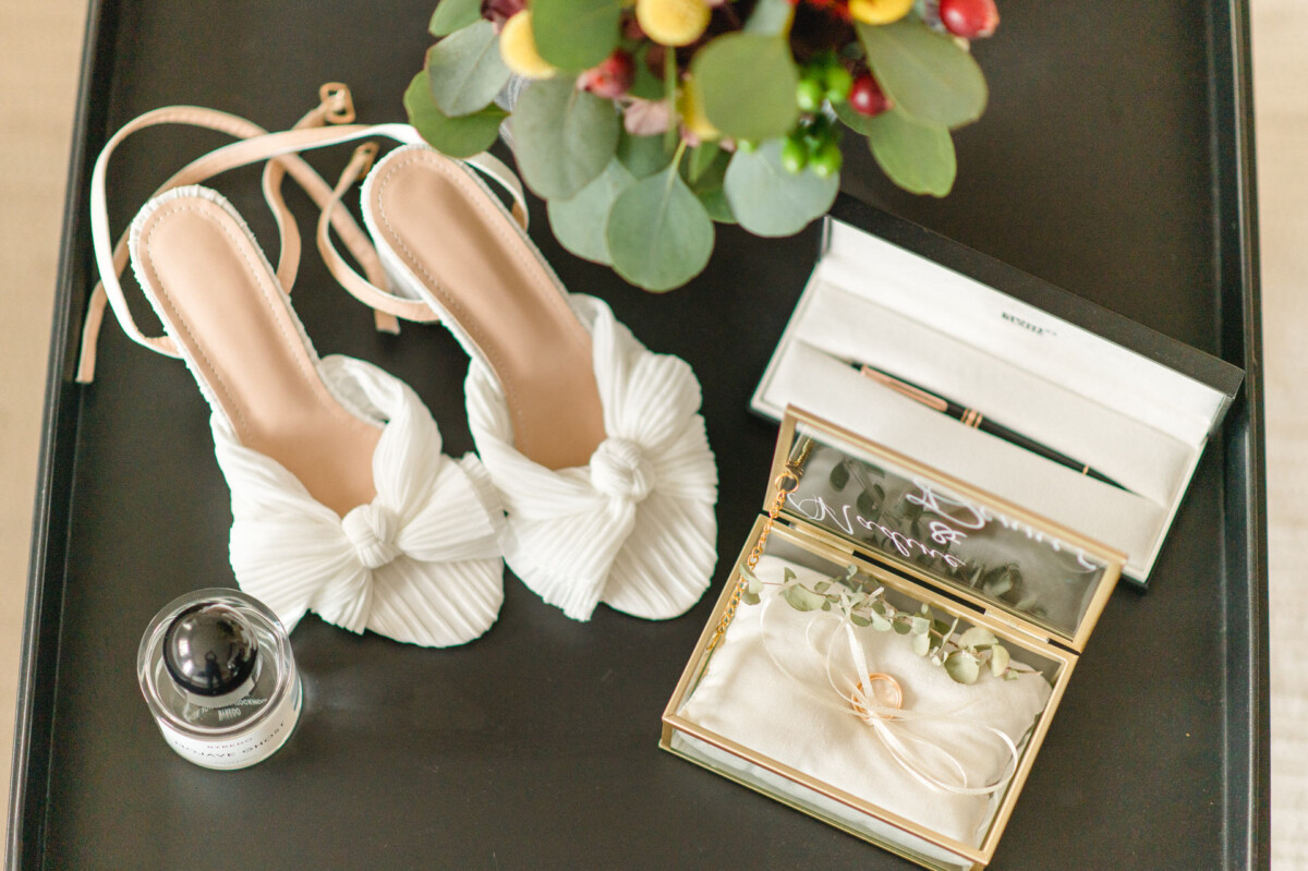 Edle Brautaccessoires auf einem schwarzen Tablett: weiße Rüschenschuhe, eine Ringschachtel mit Ringen, ein Parfümfläschchen und eine Blumendekoration.