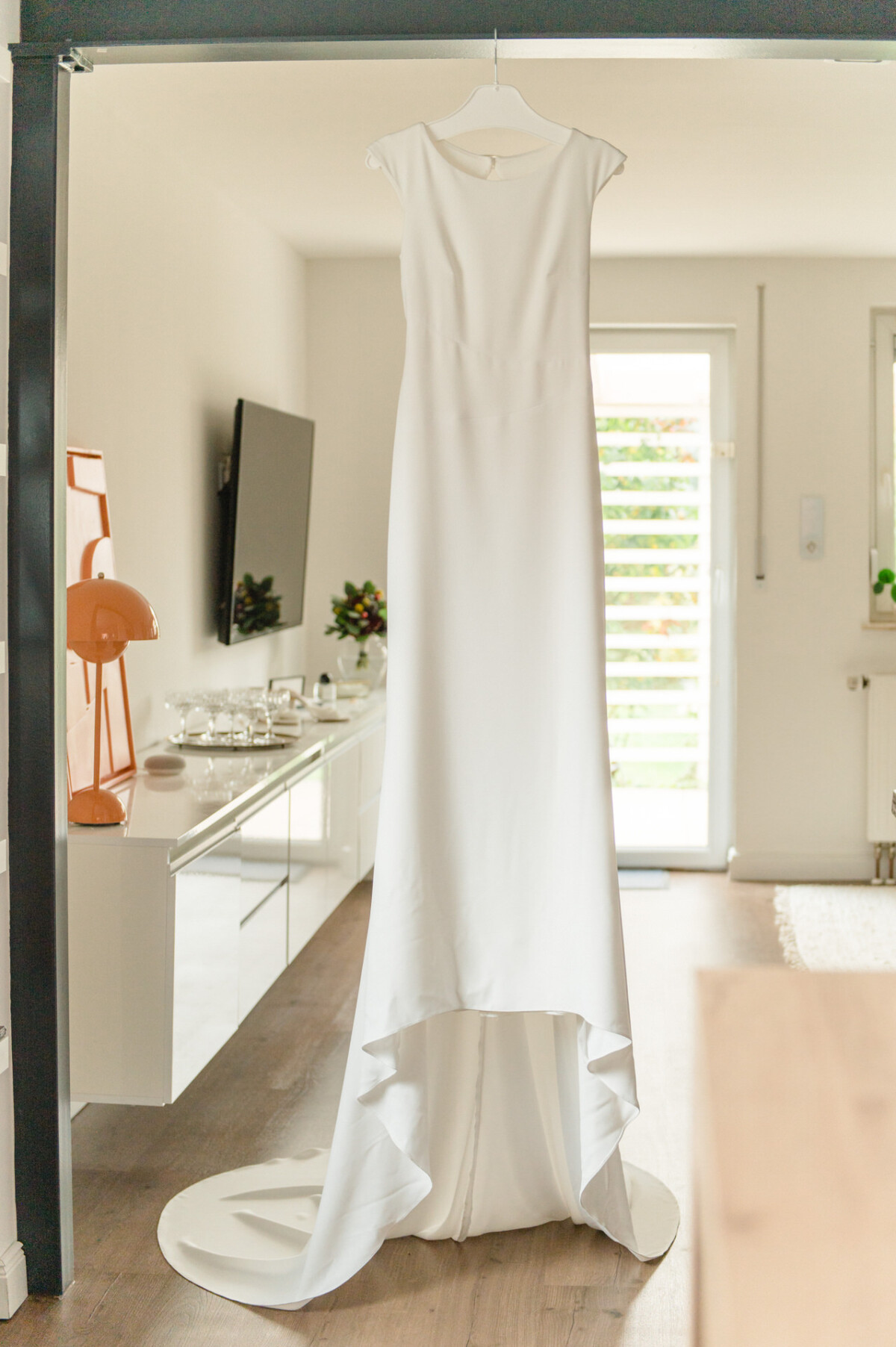 Ein weißes Brautkleid hängt in einem modernen Raum, in den helles Tageslicht durch eine Tür einfällt.