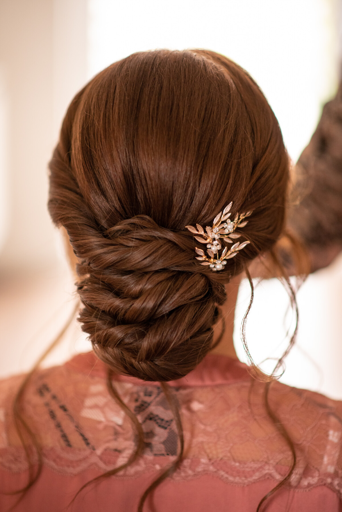 Elegante Brautfrisur, die das Haar einer Brünetten zu einem detailreichen Zopf gestylt zeigt, mit einem seitlich eingesteckten Haarteil mit Blumenmotiv, von hinten aufgenommen.