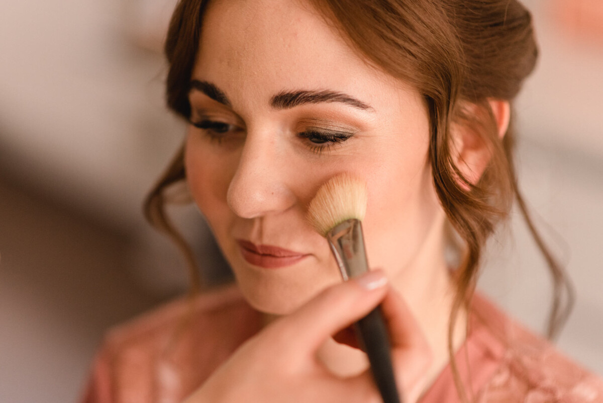 Eine Frau lässt sich mit einem Pinsel Make-up auf die Wangen auftragen und lächelt sanft, in einem Innenbereich mit warmer Beleuchtung.