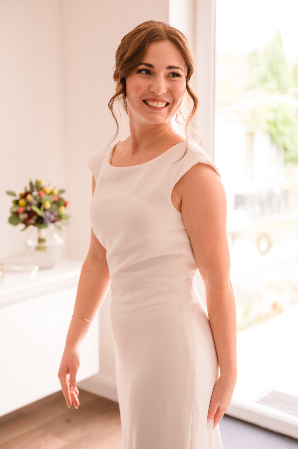 Eine lächelnde Frau in einem weißen ärmellosen Kleid steht an einem Fenster in einem gut beleuchteten Raum.