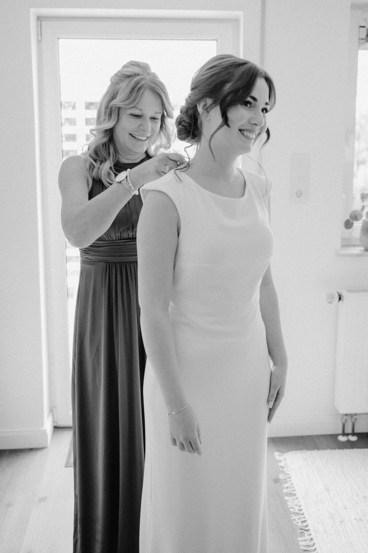 In einem hell erleuchteten Raum hilft eine Frau in einem dunklen Kleid einer lächelnden Braut im weißen Kleid mit ihrer Halskette.