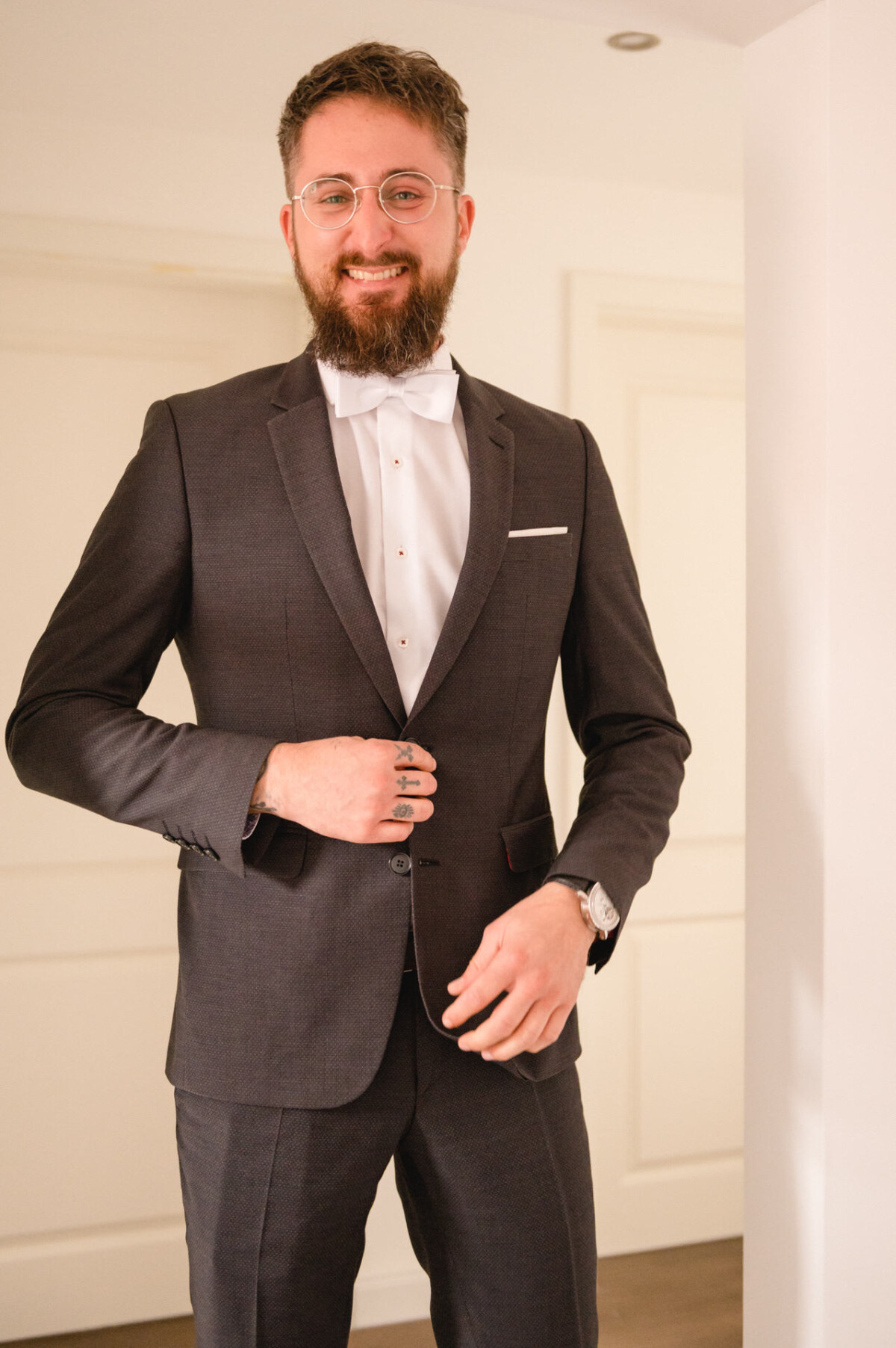 Ein bärtiger Mann in einem engen dunklen Anzug und mit Brille lächelt und rückt in einem gut beleuchteten Raum seine Manschettenknöpfe zurecht.