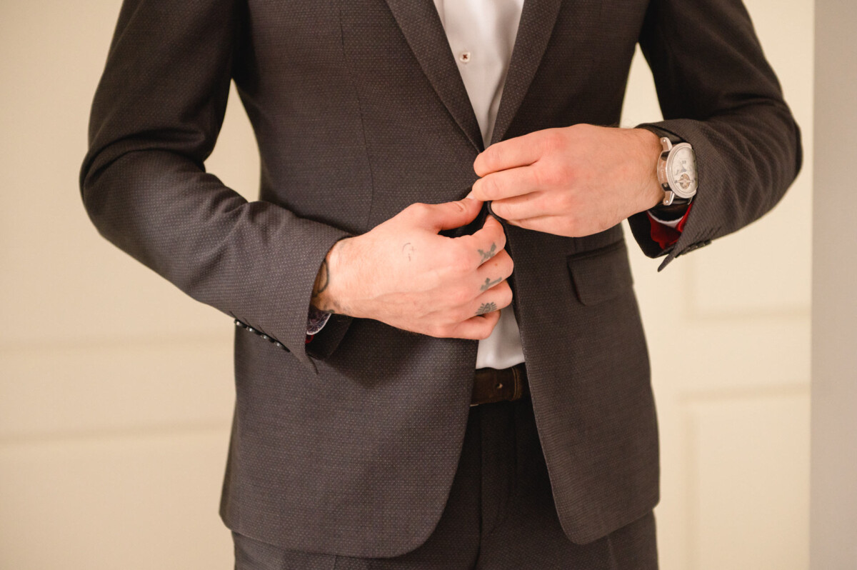 Ein Mann in dunklem Anzug und weißem Hemd richtet sein Jackett. Am linken Handgelenk trägt er eine Uhr.