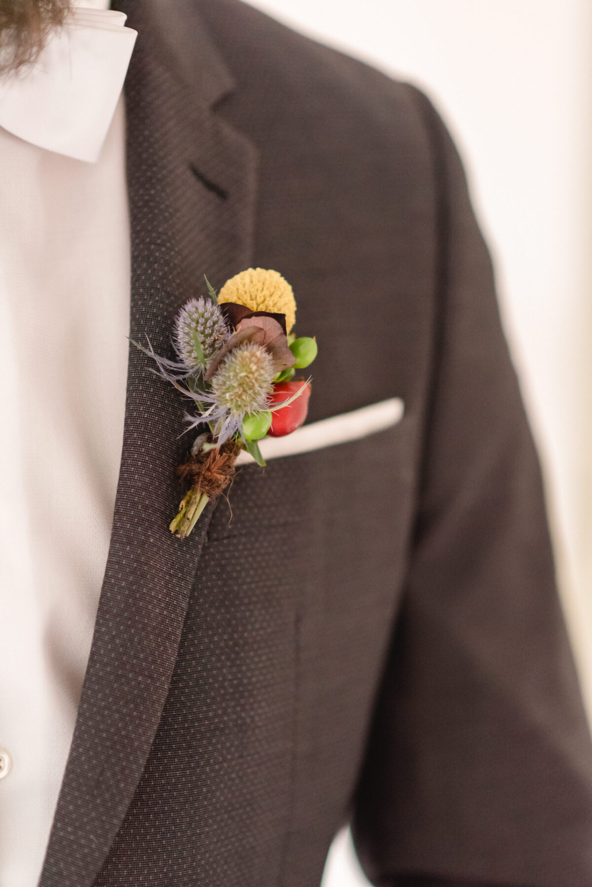 Nahaufnahme des Revers eines dunklen Anzugs des Bräutigams mit einer einzigartigen Boutonniere mit Distel, Beeren und Grünpflanzen.