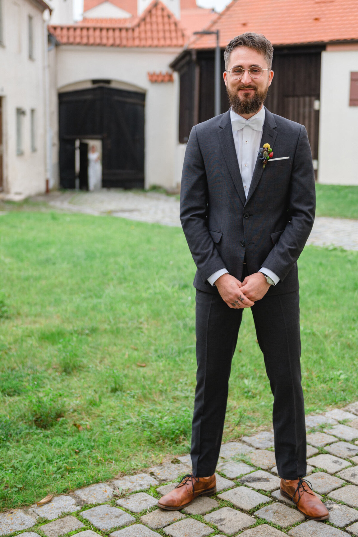 Ein Mann in dunklem Anzug und Krawatte lächelt in die Kamera und steht auf einem Kopfsteinpflasterweg mit historischen Gebäuden im Hintergrund.