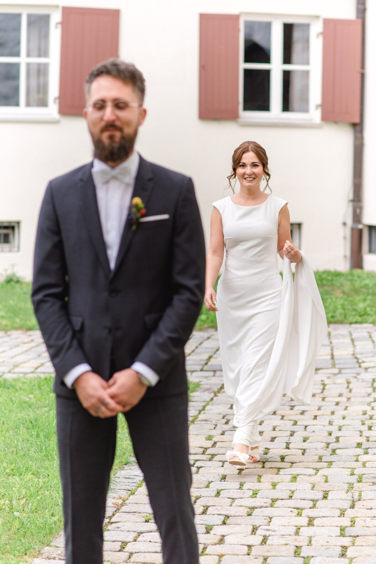Eine Braut im weißen Kleid nähert sich freudig ihrem Bräutigam, der im schwarzen Anzug auf einem Kopfsteinpflasterweg wartend steht.