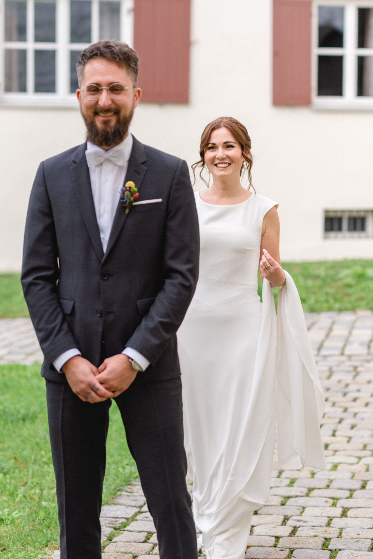 Eine Braut und ein Bräutigam gehen glücklich auf einem Kopfsteinpflasterweg spazieren. Die Braut trägt ein weißes ärmelloses Kleid und der Bräutigam einen dunklen Anzug.