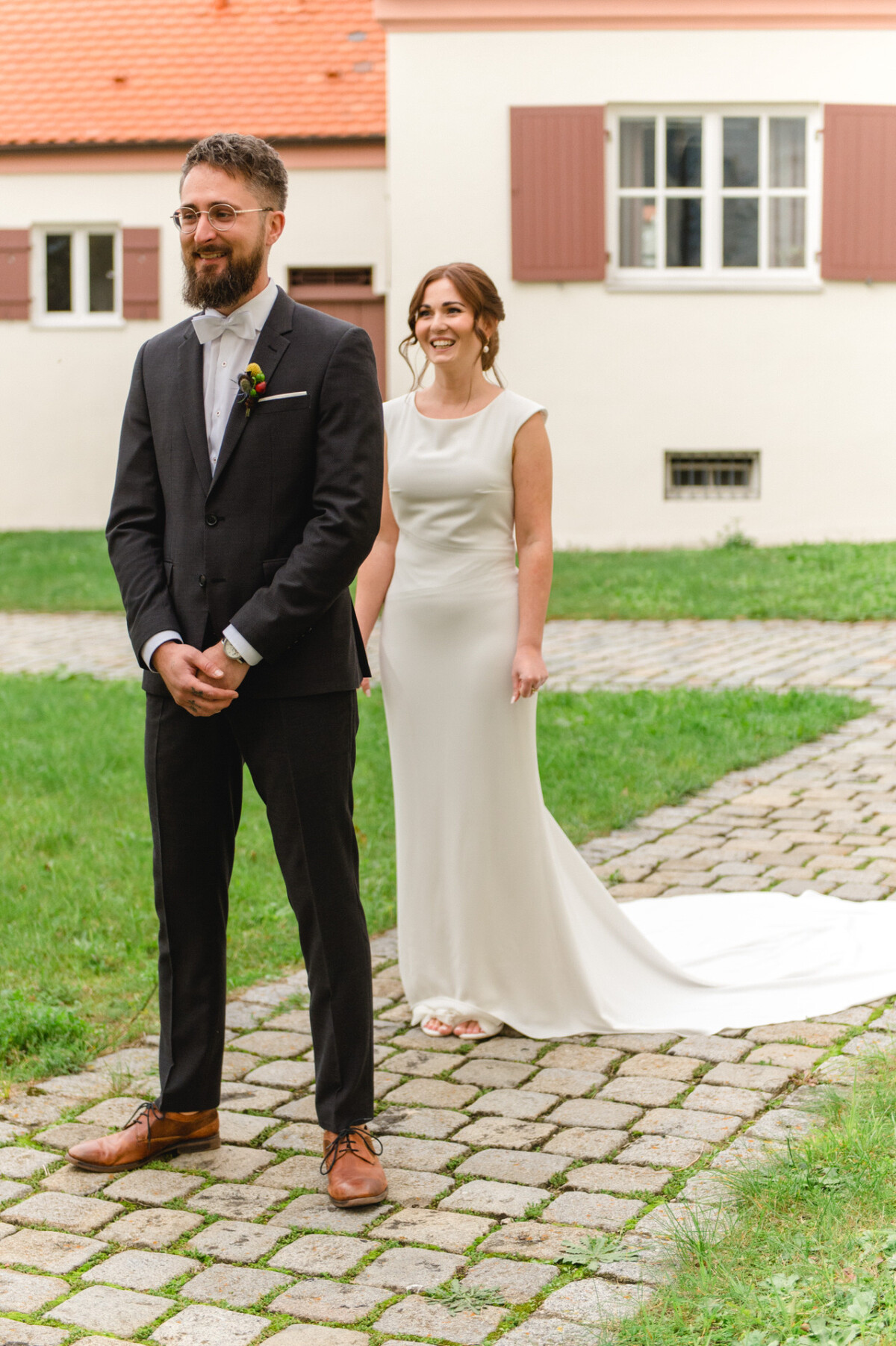 Ein Bräutigam im schwarzen Anzug und eine Braut im langen weißen Kleid stehen lächelnd auf einem Kopfsteinpflasterweg, im Hintergrund ist ein Gebäude zu sehen.
