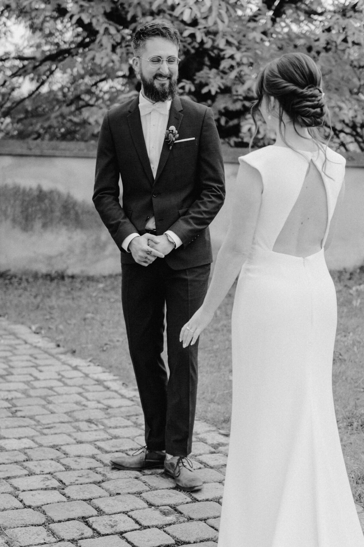 Ein Bräutigam im schwarzen Smoking lächelt die Braut an, die ein weißes Kleid mit offenem Rücken trägt, während sie auf einem Kopfsteinpflasterweg stehen.