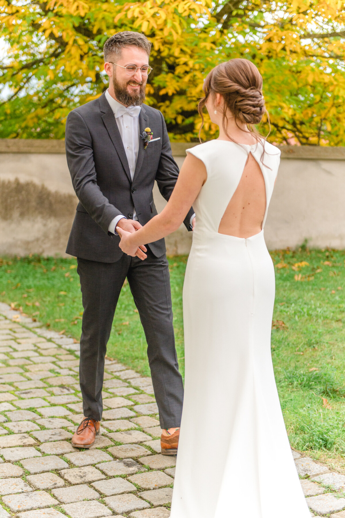 Ein Hochzeitspaar hält Händchen und lächelt sich an, die Braut trägt ein weißes rückenfreies Kleid und der Bräutigam einen grauen Anzug, im Freien mit Herbstlaub im Hintergrund.