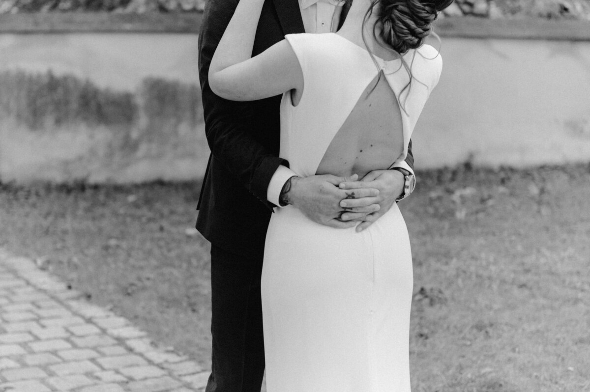 Schwarzweißfoto eines Mannes im Anzug, der eine Frau im weißen Kleid von hinten umarmt.