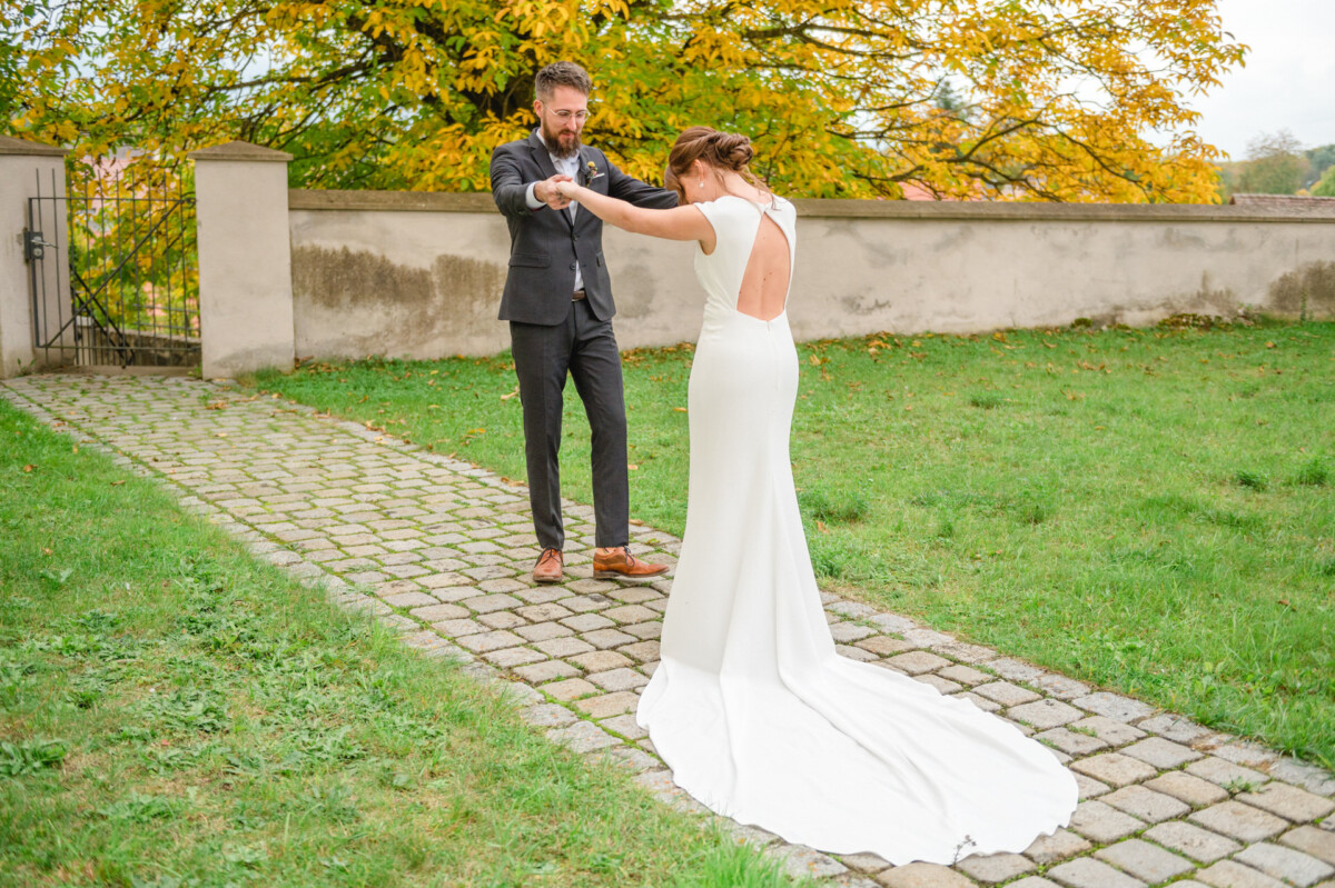 Braut und Bräutigam teilen einen spielerischen Moment, wobei der Bräutigam die Braut an der Hand durch einen Garten mit Herbstlaub im Hintergrund wirbelt.