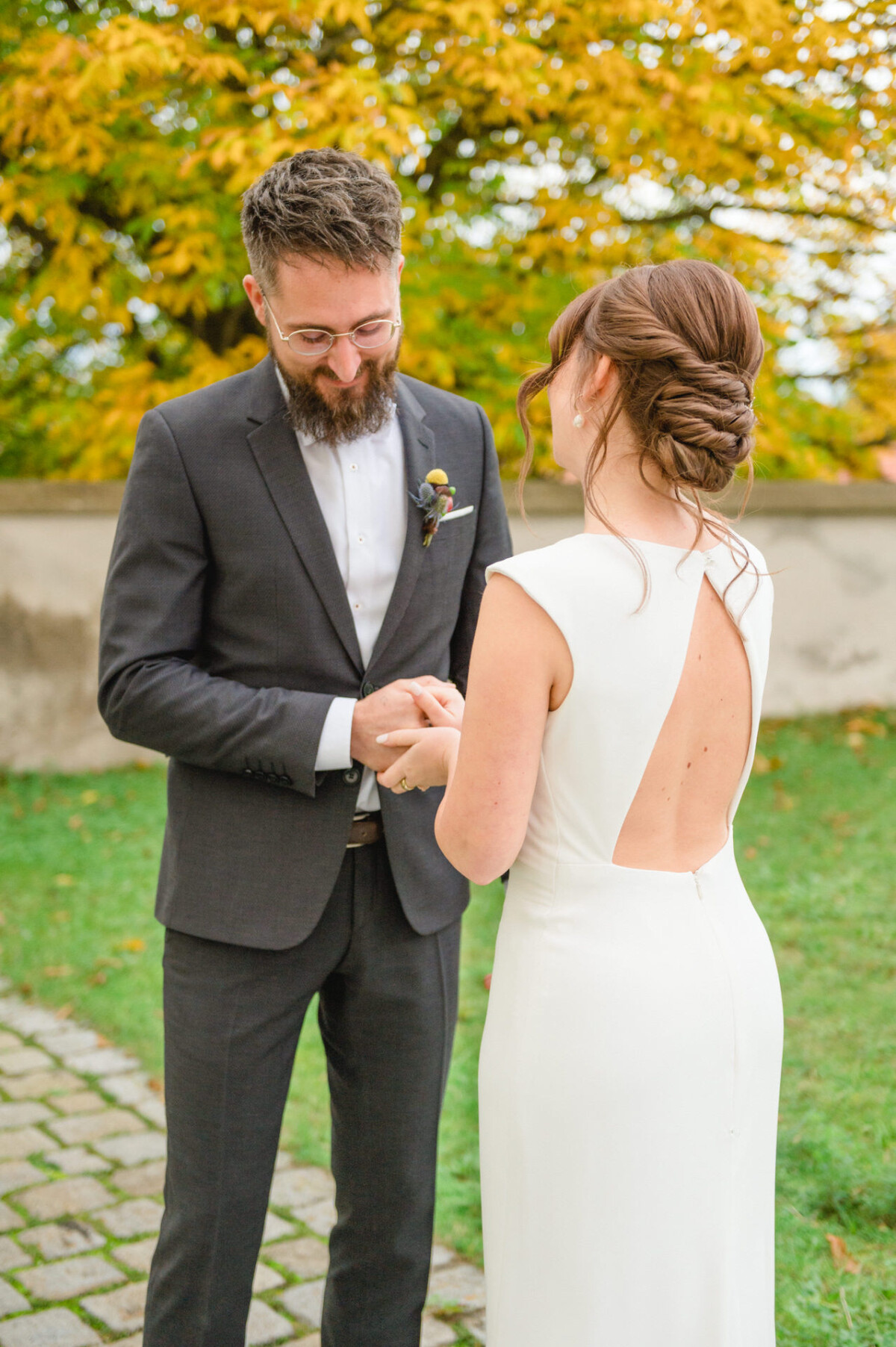 Eine Braut und ein Bräutigam halten in einem Park Händchen und lächeln einander an, im Hintergrund sind Herbstblätter zu sehen.