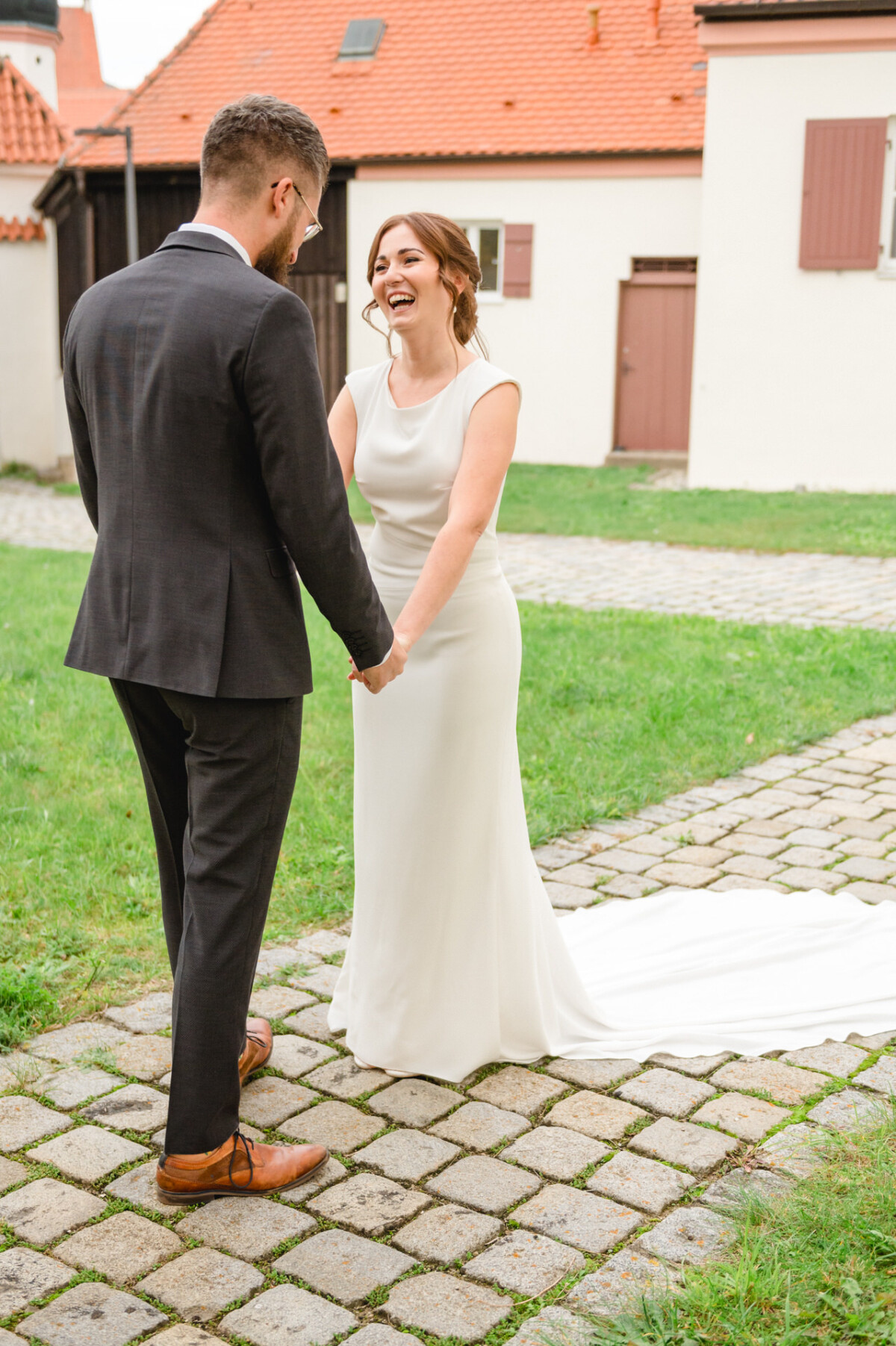 Eine fröhlich lachende Braut hält in einem Innenhof die Hand ihres Bräutigams, beide tragen Hochzeitskleidung.