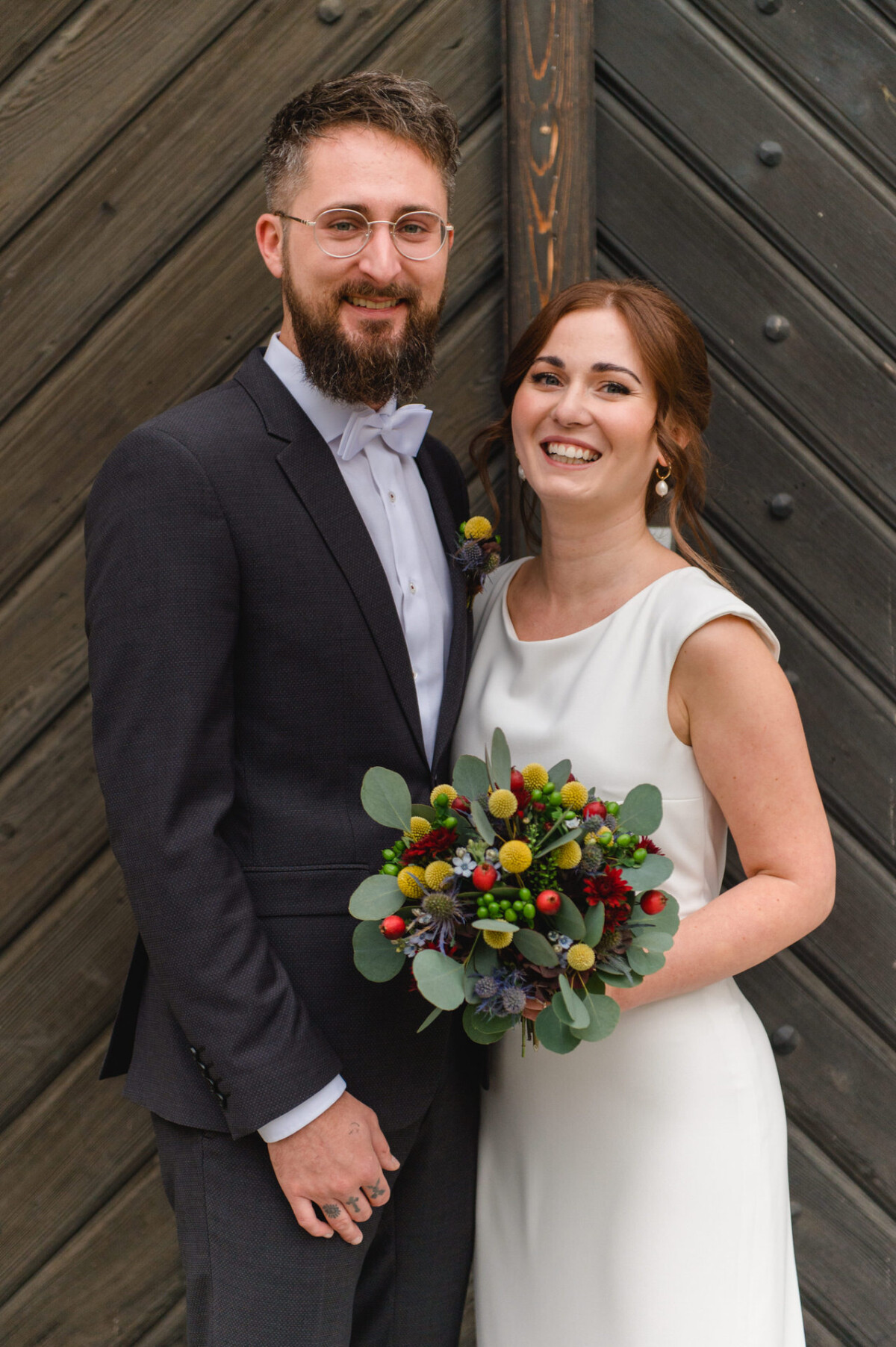 Eine Braut im weißen Kleid und ein Bräutigam im dunklen Anzug stehen lächelnd eng beieinander, wobei die Braut einen bunten Blumenstrauß in der Hand hält.