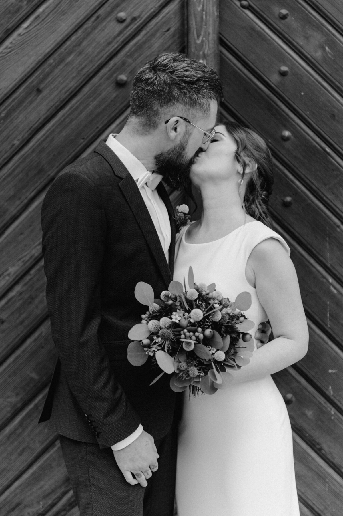 Braut und Bräutigam stehen vor einer dunklen Holztür und küssen sich zärtlich. Die Braut hält einen Strauß dunkel getönter Blumen in der Hand. Beide tragen formelle Hochzeitskleidung.