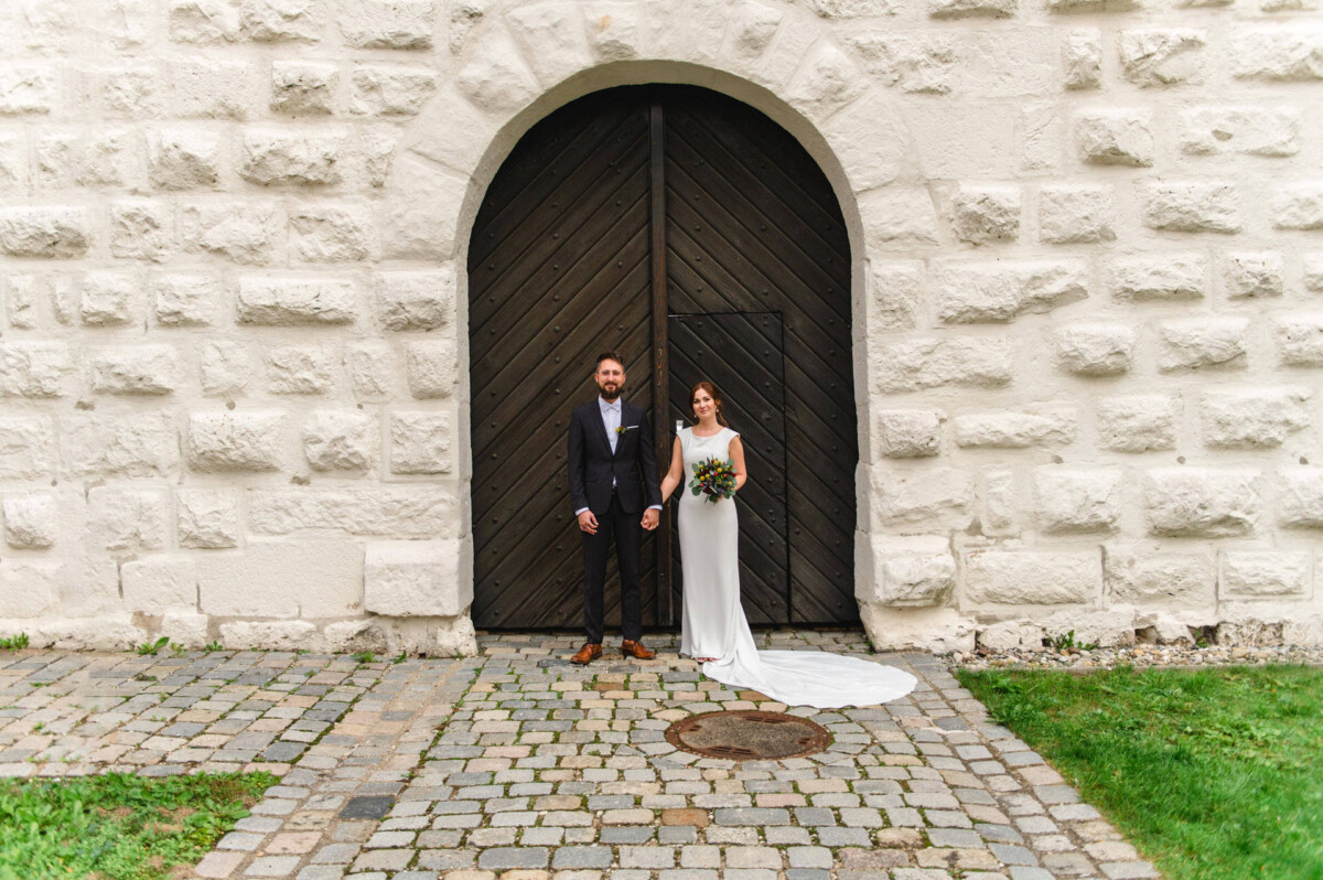Eine Braut und ein Bräutigam stehen zusammen vor einer großen, gewölbten Holztür in einer weißen Steinmauer, halten sich an den Händen und lächeln.