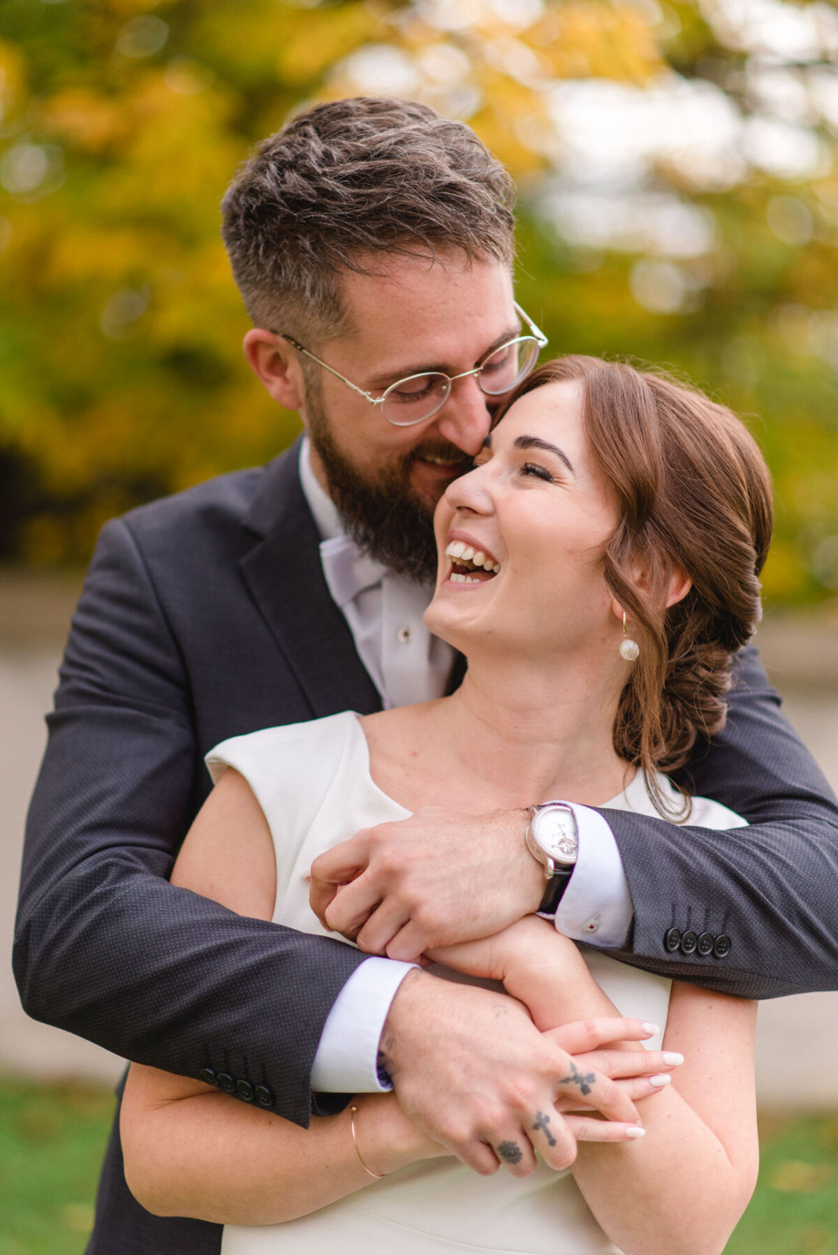 Ein fröhliches Brautpaar, das herzlich lacht, während der Bräutigam die Braut in einer herbstlichen Parklandschaft von hinten umarmt.