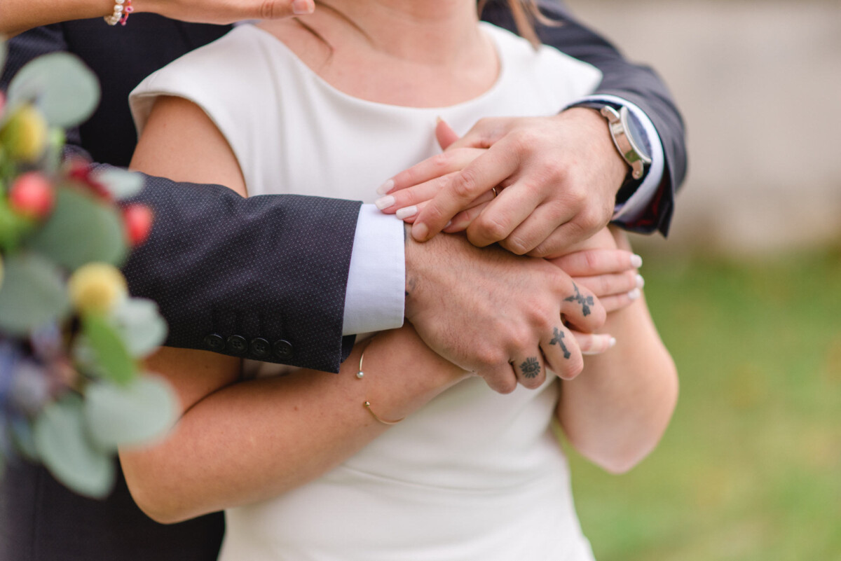 Nahaufnahme einer Braut und eines Bräutigams, die Händchen halten. Zu sehen sind die tätowierten Finger der Braut und die Uhr des Bräutigams.