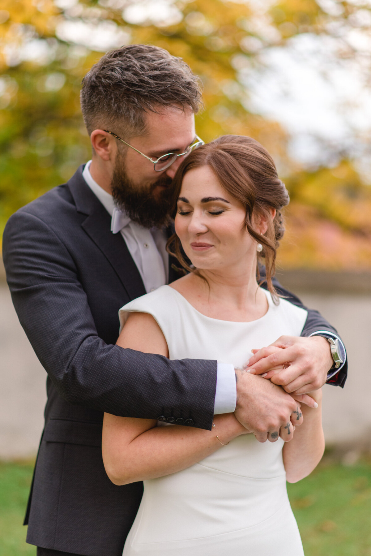 Ein bärtiger Mann im Anzug umarmt eine Frau im weißen Kleid, beide lächeln sanft vor einem herbstlichen Hintergrund.