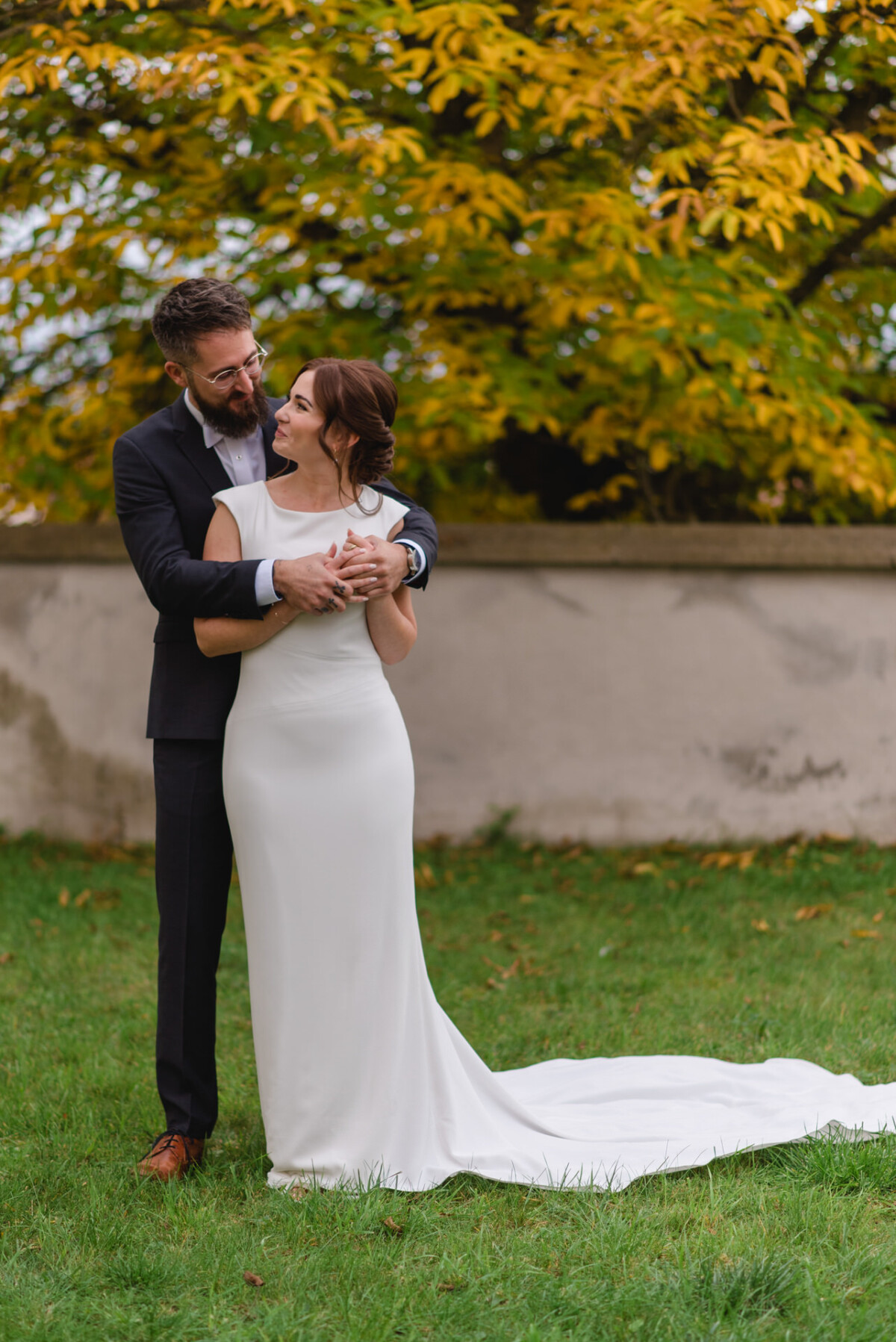 Ein frisch verheiratetes Paar umarmt sich im Freien mit Herbstblättern im Hintergrund. Der Bräutigam trägt einen Anzug und die Braut ein langes weißes Kleid.