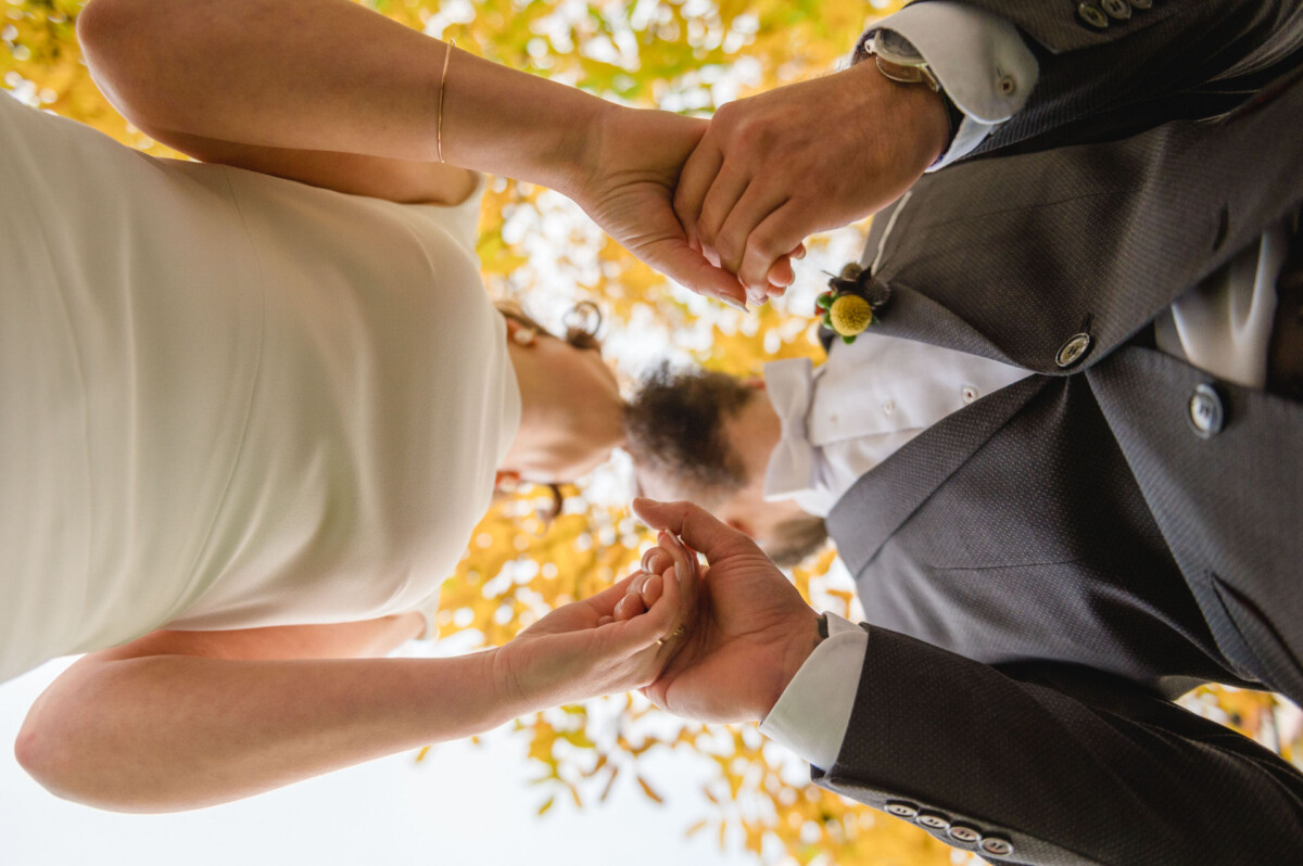 Eine Braut und ein Bräutigam halten mit einem Kind unter einem Baldachin aus Herbstblättern die Hände, von unten betrachtet.