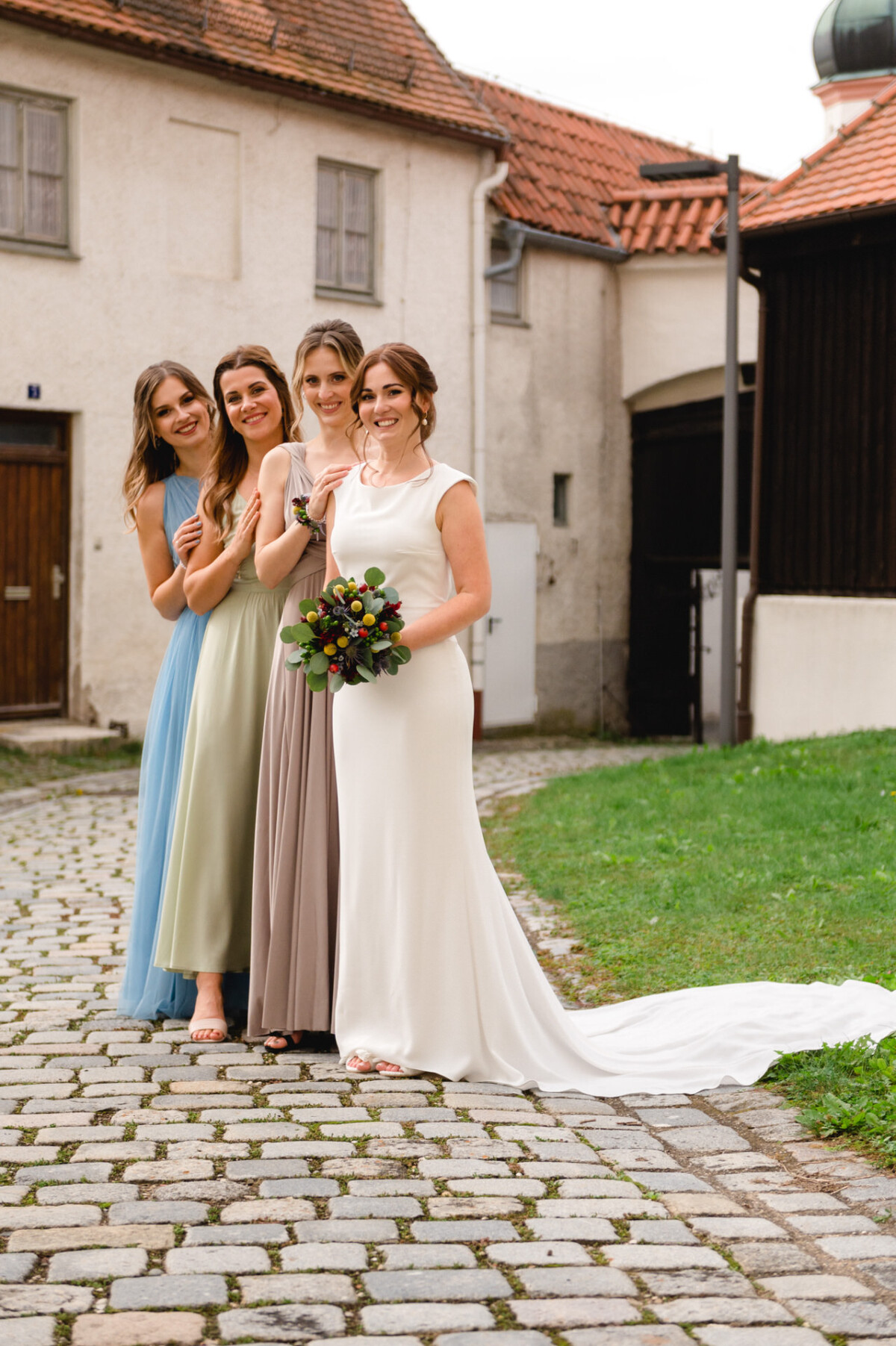 Vier Frauen in eleganten Kleidern, eine in Weiß und die anderen in Pastellfarben, lächeln und posieren gemeinsam auf einer Kopfsteinpflasterstraße.