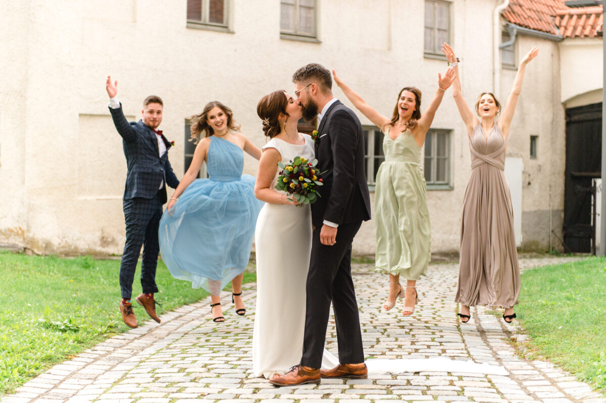Eine fröhliche Hochzeitsszene mit einem sich küssenden Paar und vier Freunden, die um sie herum in einem Innenhof feiern.