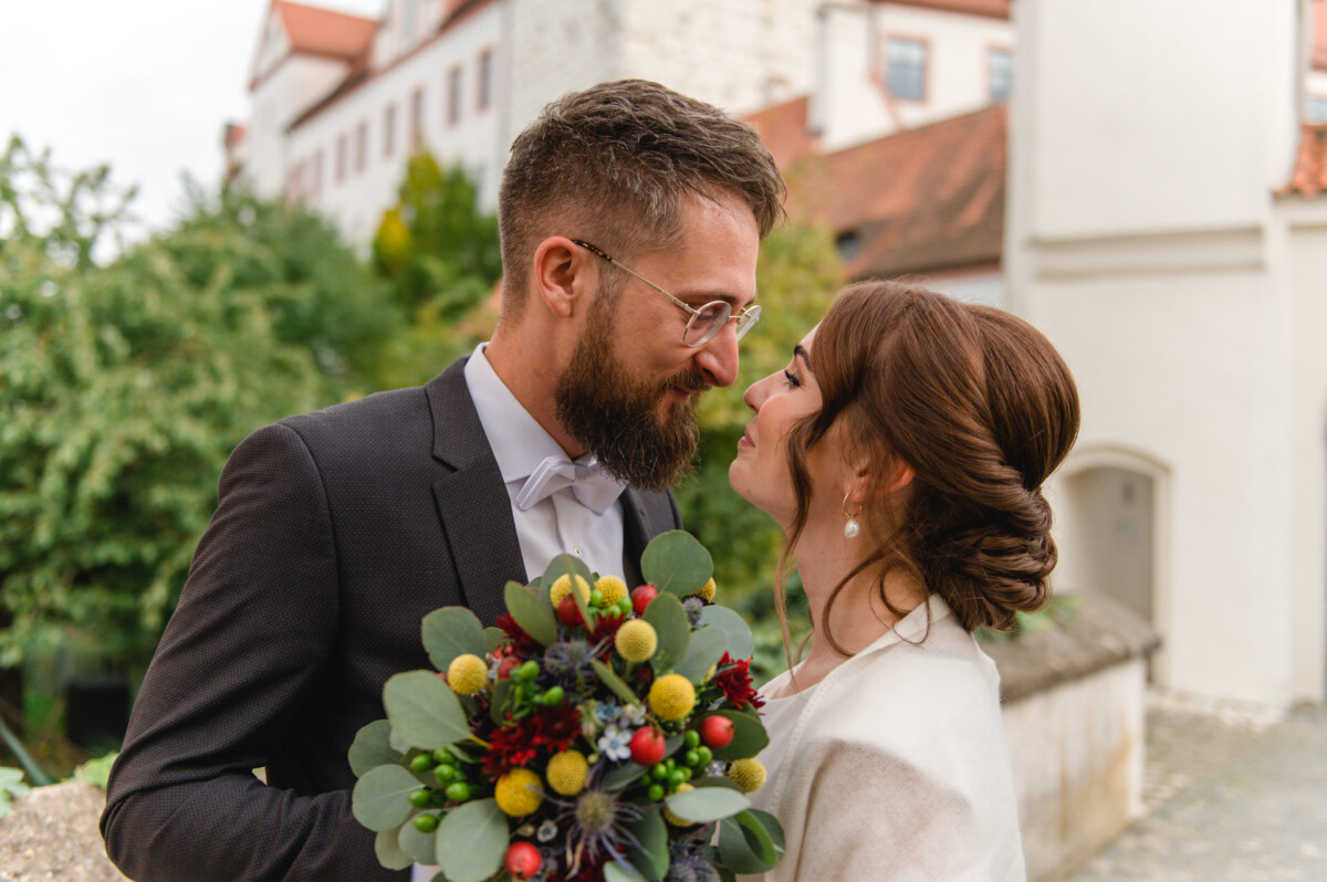 Eine Braut und ein Bräutigam, die sich gerade küssen, halten einen Blumenstrauß in der Hand, im Hintergrund ist ein altes Gebäude zu sehen.