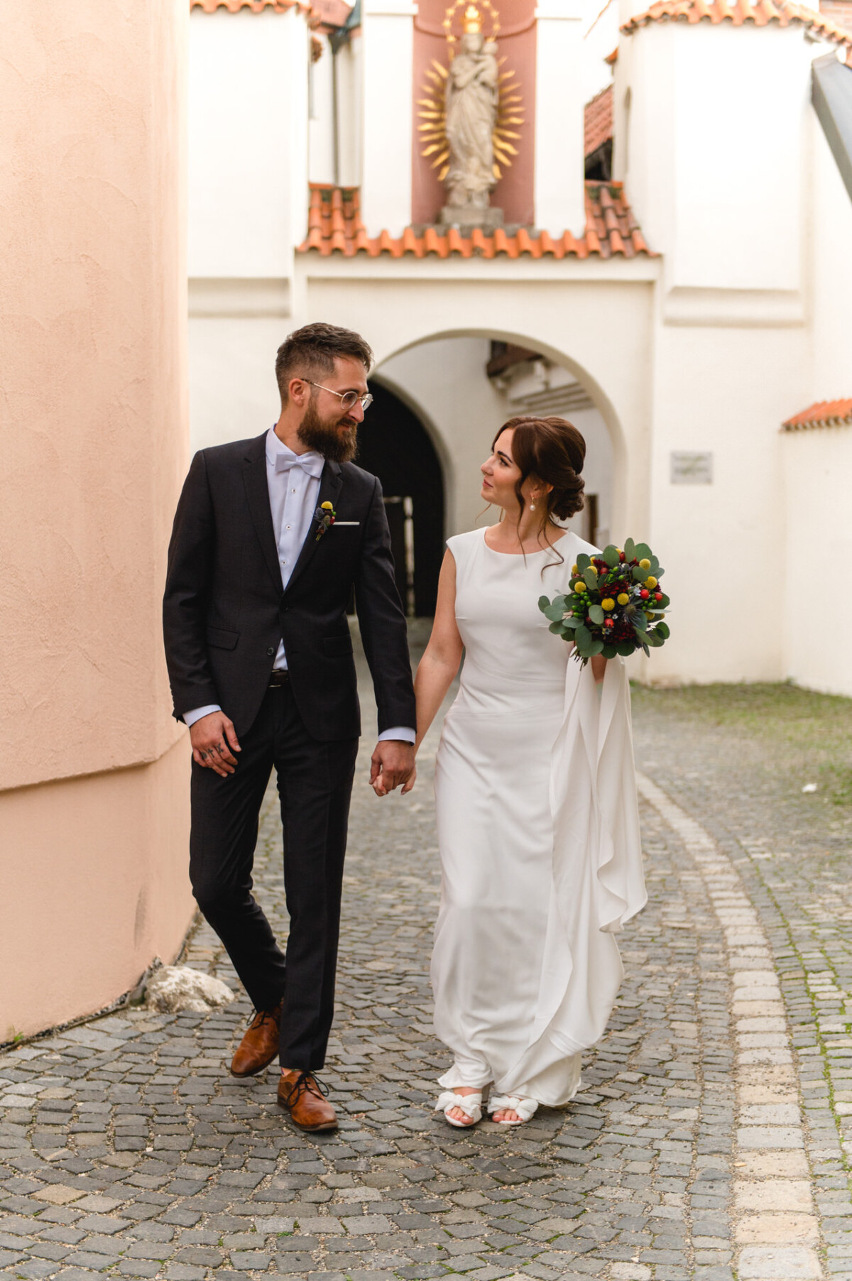 Eine Braut und ein Bräutigam gehen Hand in Hand einen Kopfsteinpflasterweg entlang und lächeln einander an. Die Braut hält einen Strauß dunkler Blumen in der Hand.