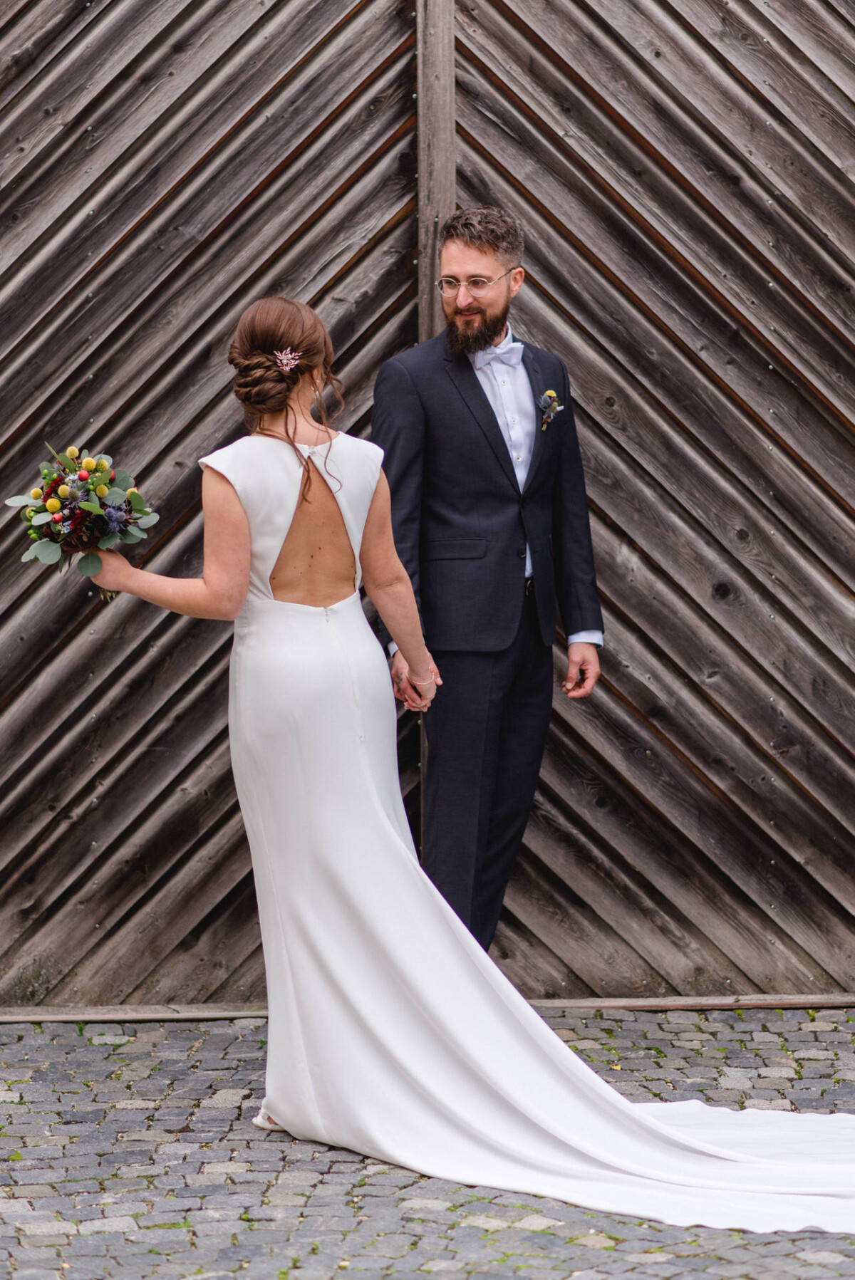 Eine Braut in einem weißen Kleid mit langer Schleppe, die einen Blumenstrauß hält, und ein Bräutigam in einem dunklen Anzug, beide stehen auf einem Kopfsteinpflasterboden vor einer hölzernen Kulisse.