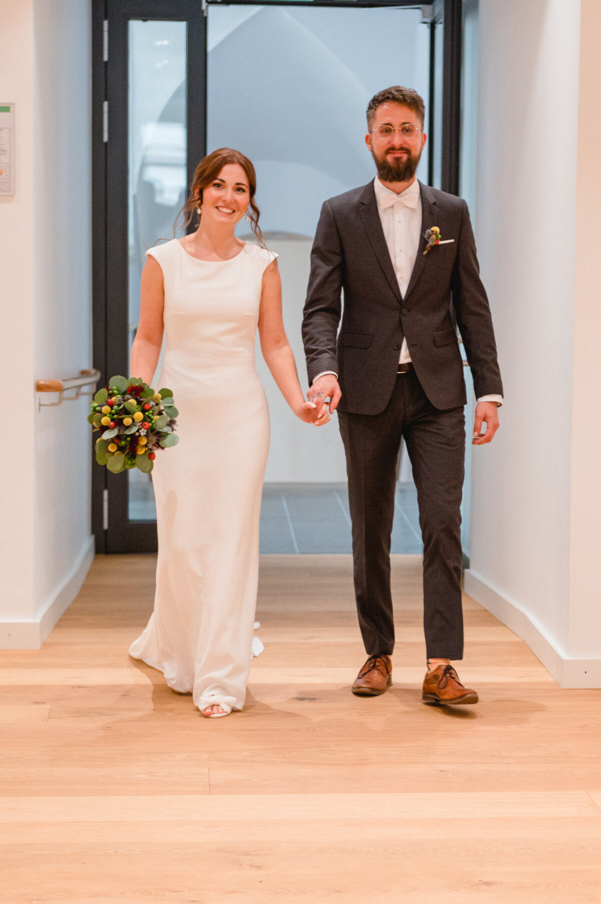 Eine Braut in einem weißen Kleid und ein Bräutigam in einem dunklen Anzug halten Händchen und lächeln, während sie durch einen Flur gehen.