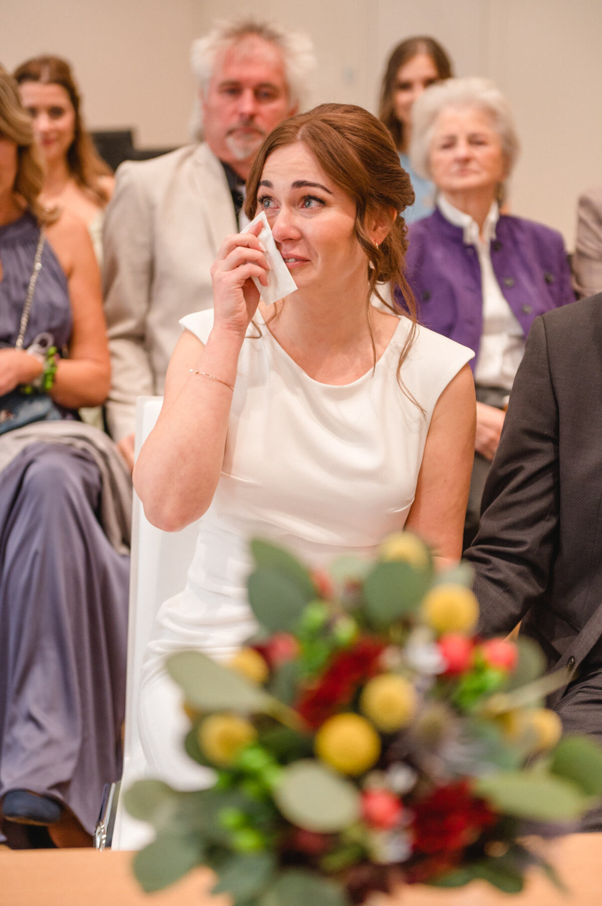 Eine Braut im weißen Kleid wischt sich während einer Hochzeitszeremonie Tränen aus den Augen, um sie herum sitzen Gäste.