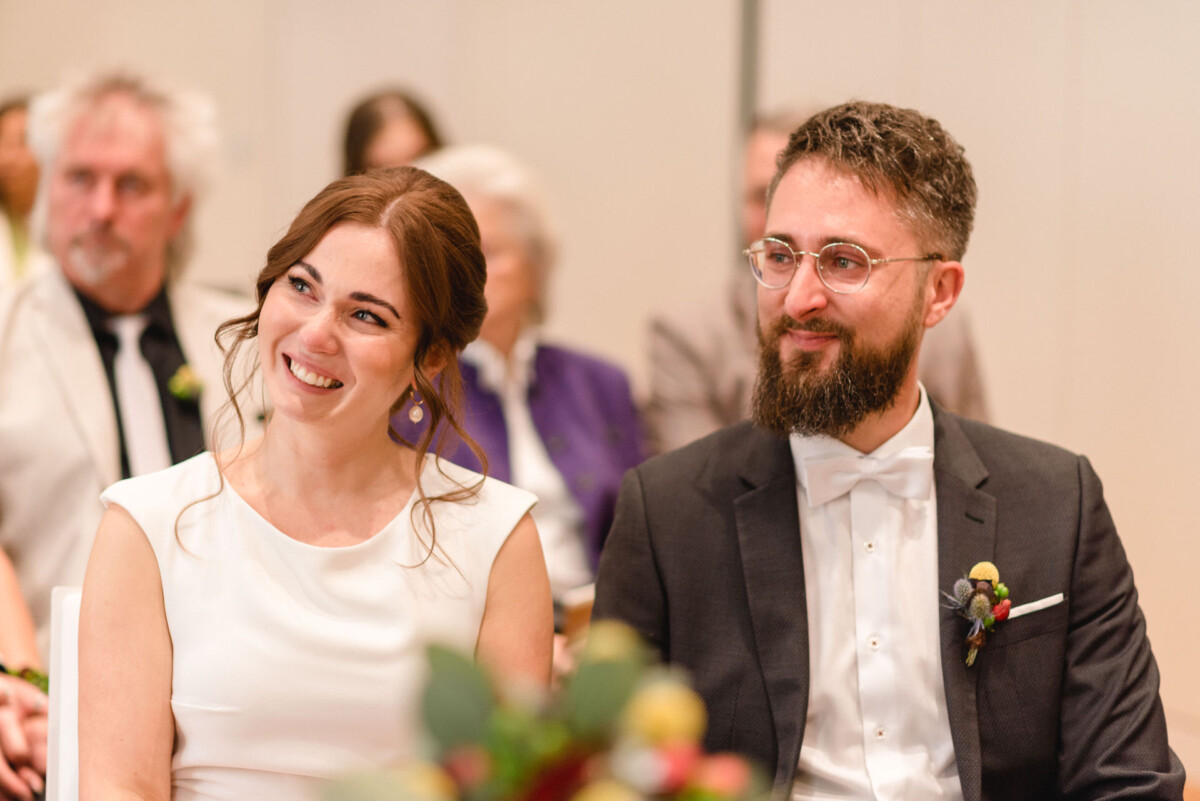 Eine Braut und ein Bräutigam lächeln und blicken aufmerksam bei einer Hochzeitszeremonie, elegant gekleidet in einem weißen Kleid und einem schwarzen Smoking.